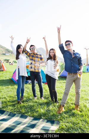 Smiling Chinese male and female friends raising arms and making hand gestures having fun while standing with cups of beer on concert ground at musi... Stock Photo