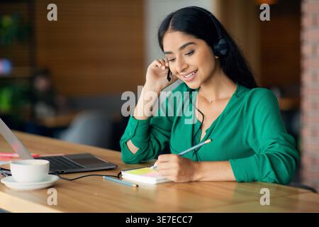 Indian woman using headset writing notes sitting at table with laptop in cafe Stock Photo