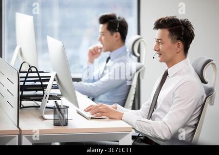 Smiling Chinese male support agents wearing headsets answering customer calls and working on computers while sitting at table in office Stock Photo