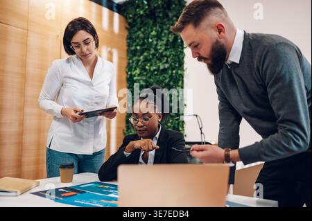Serious Black business woman in eyeglasses sitting at her desk with document reports having meeting with caucasian male and female colleagues in of... Stock Photo