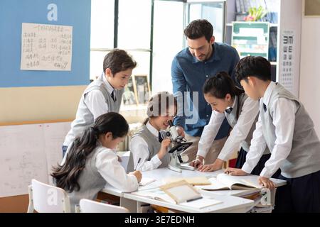 Diverse group of students using microscope with caucasian male teacher during science class in classroom Stock Photo