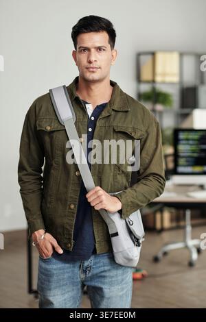 Indian male web developer with cross-body bag looking away standing in office Stock Photo