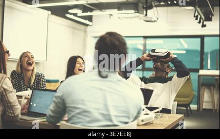 Smiling Black male developer with braids wearing VR headset and experiencing virtual reality while sitting at table with diverse colleagues laughin... Stock Photo
