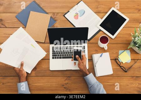 Black businessman reviewing documents and using smartphone while sitting at wooden desk with laptop and various objects in office Stock Photo