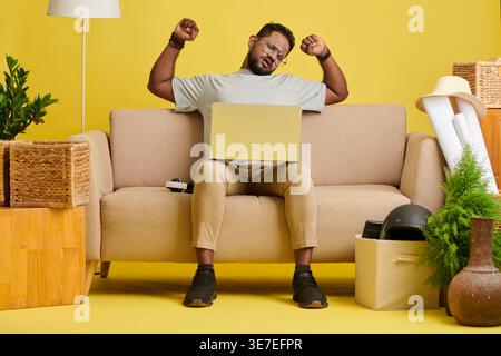 Asian man with closed eyes wearing eyeglasses stretching arms sitting with laptop on sofa in living room at home Stock Photo