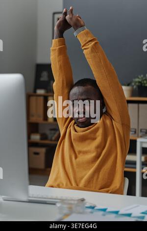 Tired Black man stretching arms looking at computer screen sitting at table in office Stock Photo