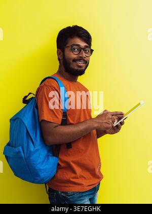 Smiling Indian man in orange shirt with eyeglasses and blue backpack standing while using tablet against yellow background in studio Stock Photo