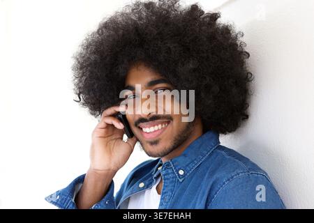 Smiling Black man with afro hairstyle having phone call standing leaning on white wall Stock Photo
