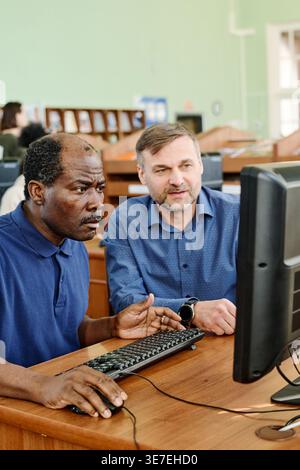 Middle-aged Black male immigrant sitting at desk with caucasian male instructor helping him to understand task on computer in library Stock Photo