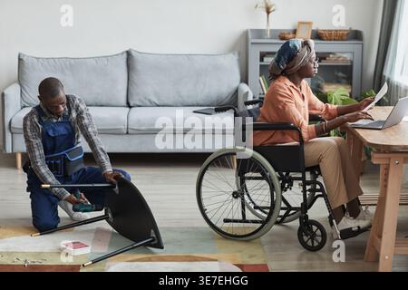 Disabled Black woman wearing head wrap and eyeglasses working on laptop while sitting in wheelchair near handyman assembling furniture in living ro... Stock Photo