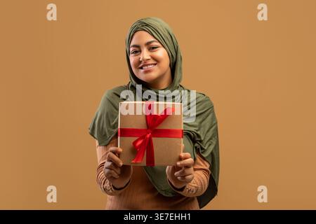 Smiling Middle Eastern woman wearing hijab holding gift box while standing against brown background in studio Stock Photo