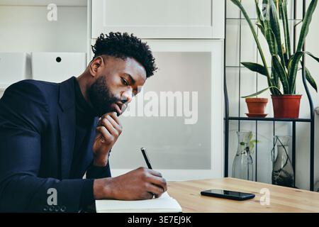 Black businessman with beard writing in notebook and leaning chin on hand thinking while sitting at desk with smartphone in office Stock Photo