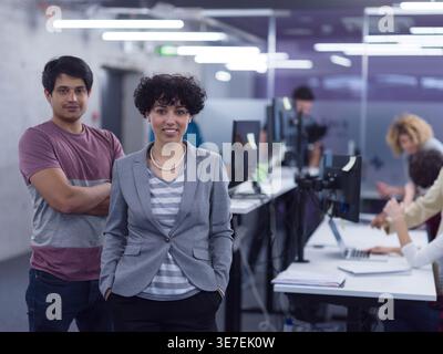 Smiling Mixed race woman with hands in pockets standing near Mixed race man with arms crossed in office Stock Photo