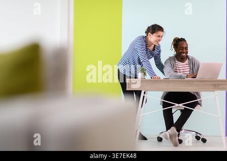 Smiling Black businessman with braids sitting at table working on laptop and having discussion with caucasian female colleague standing beside him ... Stock Photo