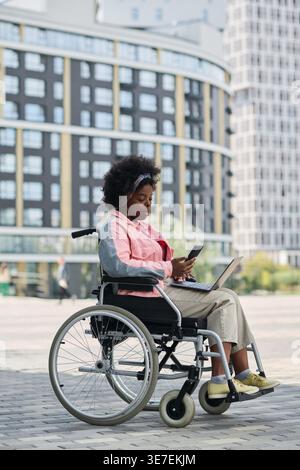 Disabled Black woman in wheelchair using smartphone in city street during daytime Stock Photo