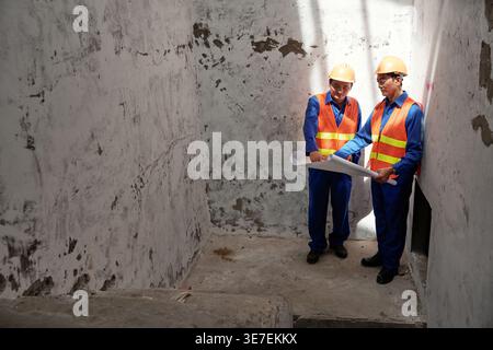 Asian male construction workers discussing over building plan while standing inside building in construction site Stock Photo