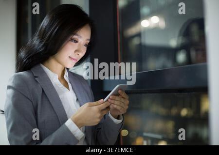 Smiling Asian business woman using smartphone while standing near window in office at night Stock Photo