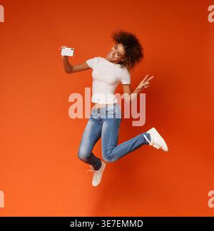 Blissful Black woman jumping and taking peace selfie on smartphone against orange background in studio Stock Photo