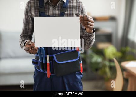 Cropped image of Black handyman with tool belt holding blank white sign standing at home Stock Photo