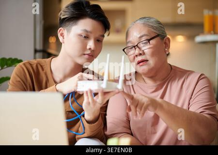 Asian grandmother wearing eyeglasses learning how to setup wifi router with her grandson while sitting on sofa at home Stock Photo