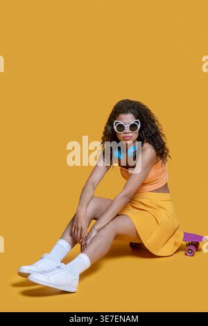 African American female leaning arms on knees sitting on skateboard against wall Stock Photo