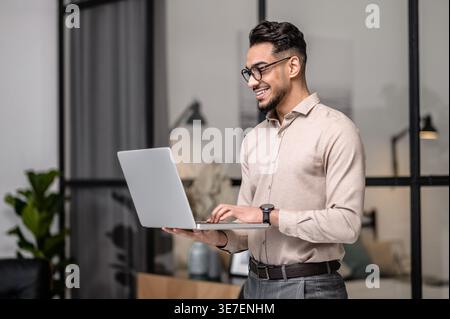 Smiling Arab man wearing eyeglasses having video call on laptop while standing at home Stock Photo