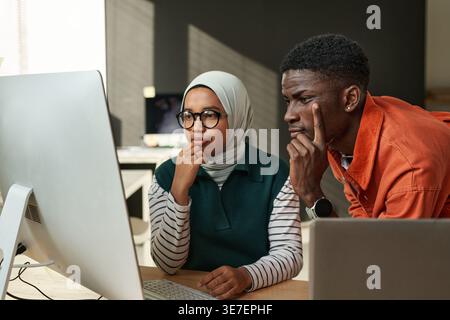 Black male and Mixed race Muslim female software developers working together on a project staring at computer monitor sitting at desk in office Stock Photo
