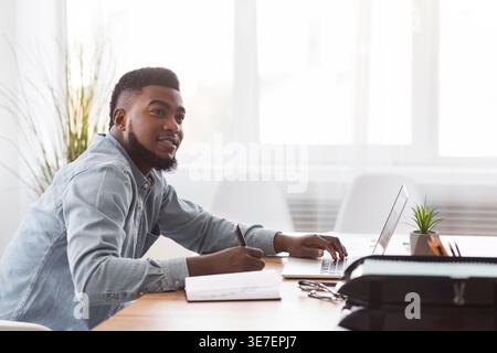 Smiling Black businessman looking away taking notes from laptop sitting at table in office Stock Photo