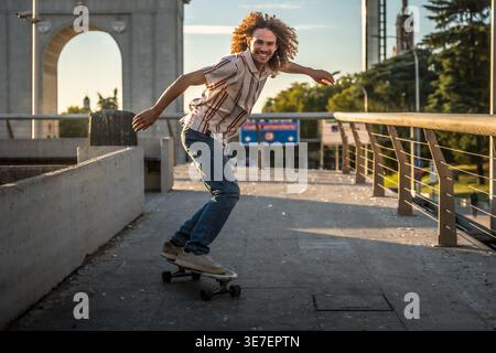 Smiling Hispanic man with curly hair riding on skateboard in city street on sunny day Stock Photo
