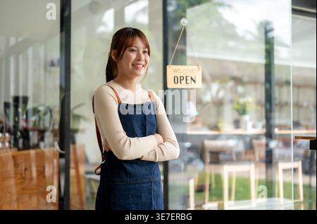 Smiling Asian woman with crossed arms looking away standing near entrance door in coffee shop Stock Photo