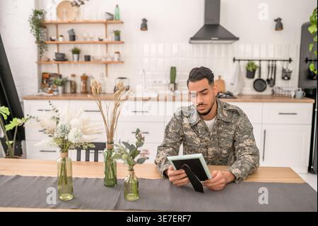 Arab military man with sad facial expression wearing camouflage uniform holding and looking at picture frame while sitting at kitchen table at home Stock Photo