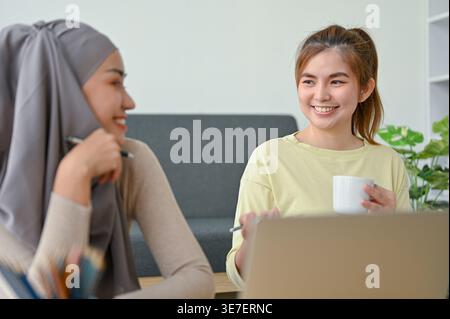 Smiling Asian female friends talking at each other while sitting at table with laptop at home Stock Photo