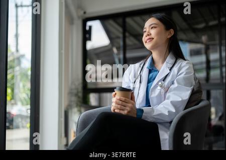 Smiling Asian female doctor looking away holding take away coffee sitting on chair in hospital office Stock Photo