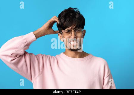 Middle Eastern man wearing eyeglasses and pink sweatshirt scratching his head while standing against blue background in studio Stock Photo
