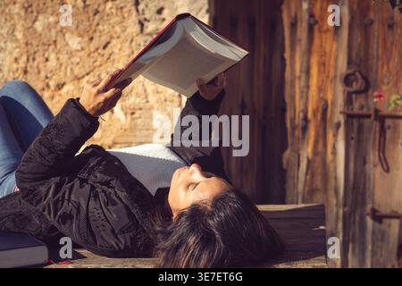 Middle-aged Hispanic woman reading book while lying on wooden table in the garden on sunny day Stock Photo