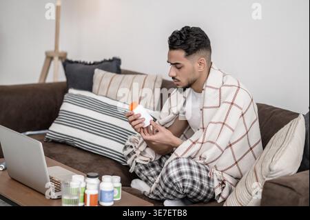 Sick Arab man wrapped in blanket holding medicine bottle sitting on sofa near table with laptop at home Stock Photo