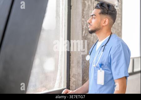 Serious Arab male doctor with stethoscope on neck looking away standing near window at home Stock Photo