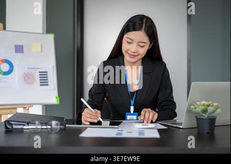 Smiling Asian business woman using tablet and calculator in analyzing financial data on reports while sitting at her desk with laptop in the office Stock Photo