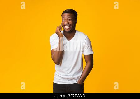 Afro Man Talking On Phone And Laughing Standing Over Yellow Background In Studio. Free Space For Text Stock Photo