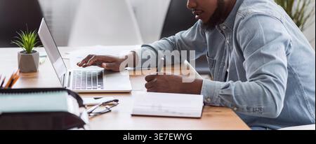 Black businessman taking notes from laptop sitting at table in office Stock Photo