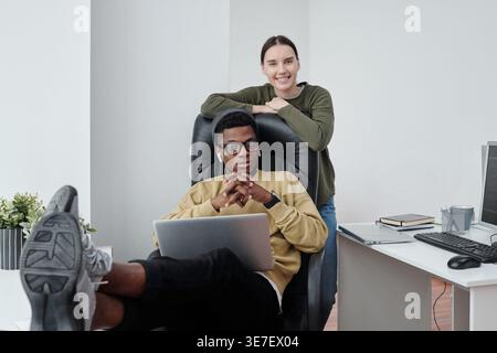 Cheerful Mixed race woman standing leaning on chair of Black man colleague with laptop sitting while foot up on table in office Stock Photo