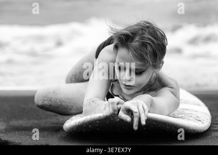 Artistic black and white portrait of pensive woman resting on surfboard on sandy beach. Sand covers her skin. The mood is melancholic and introspectiv Stock Photo