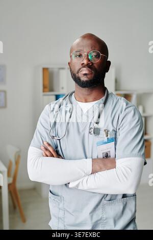 Serious Black male doctor in scrub suit with stethoscope around neck standing with arms crossed in clinic Stock Photo