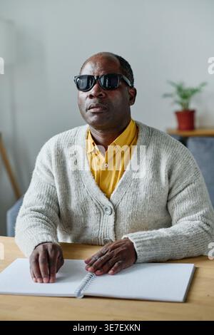 Black blind man with sunglasses sitting at the table reading braille at home Stock Photo