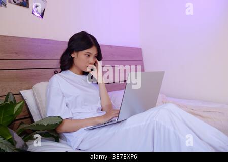 Serious Asian woman with hand on face using laptop sitting on bed at home Stock Photo