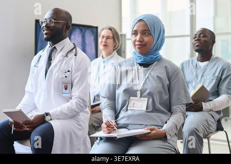Group of doctors making notes and listening to speaker while sitting on chairs during medical conference in hospital Stock Photo