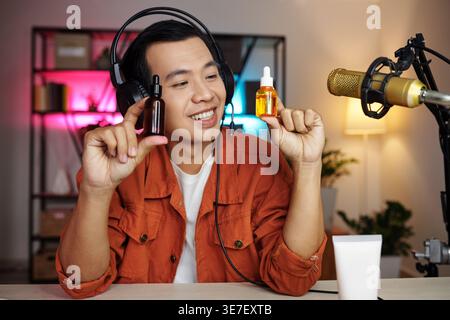 Smiling Asian male online seller wearing headset showing face serums sitting at table streaming live from home studio Stock Photo
