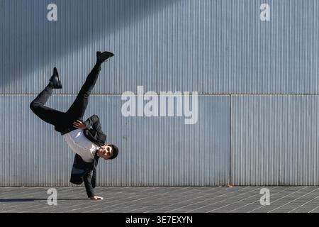 Smiling Asian businessman wearing suit doing handstand against wall on city street on sunny day Stock Photo