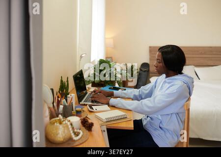 Black female student typing on her laptop sitting at study table near bed inside her dorm room during daytime Stock Photo