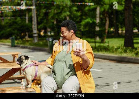Black middle-aged woman with short afro taking photos with her dog sitting on wooden bench on sunny day in park Stock Photo
